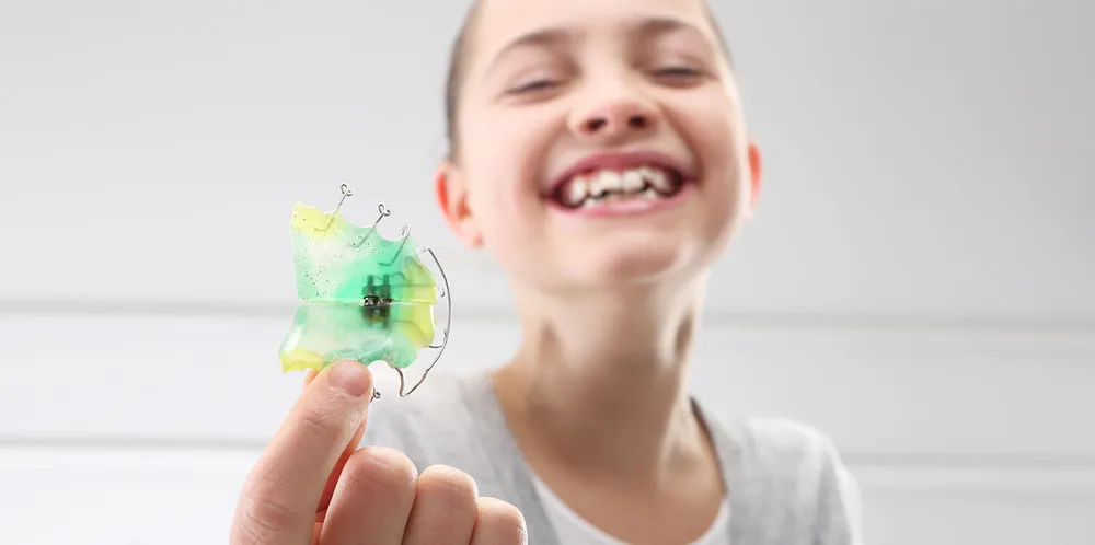 A smiling girl holding up a green and yellow removable retainer