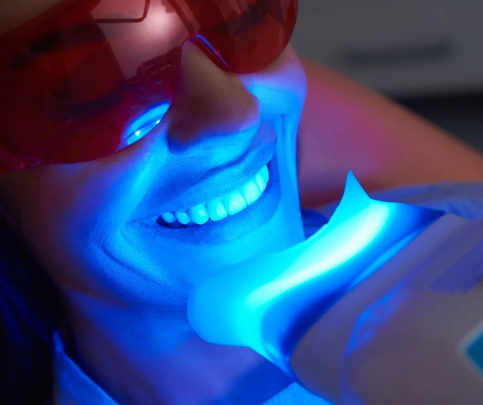 A smiling woman in protective glasses with a blue whitening light over her teeth