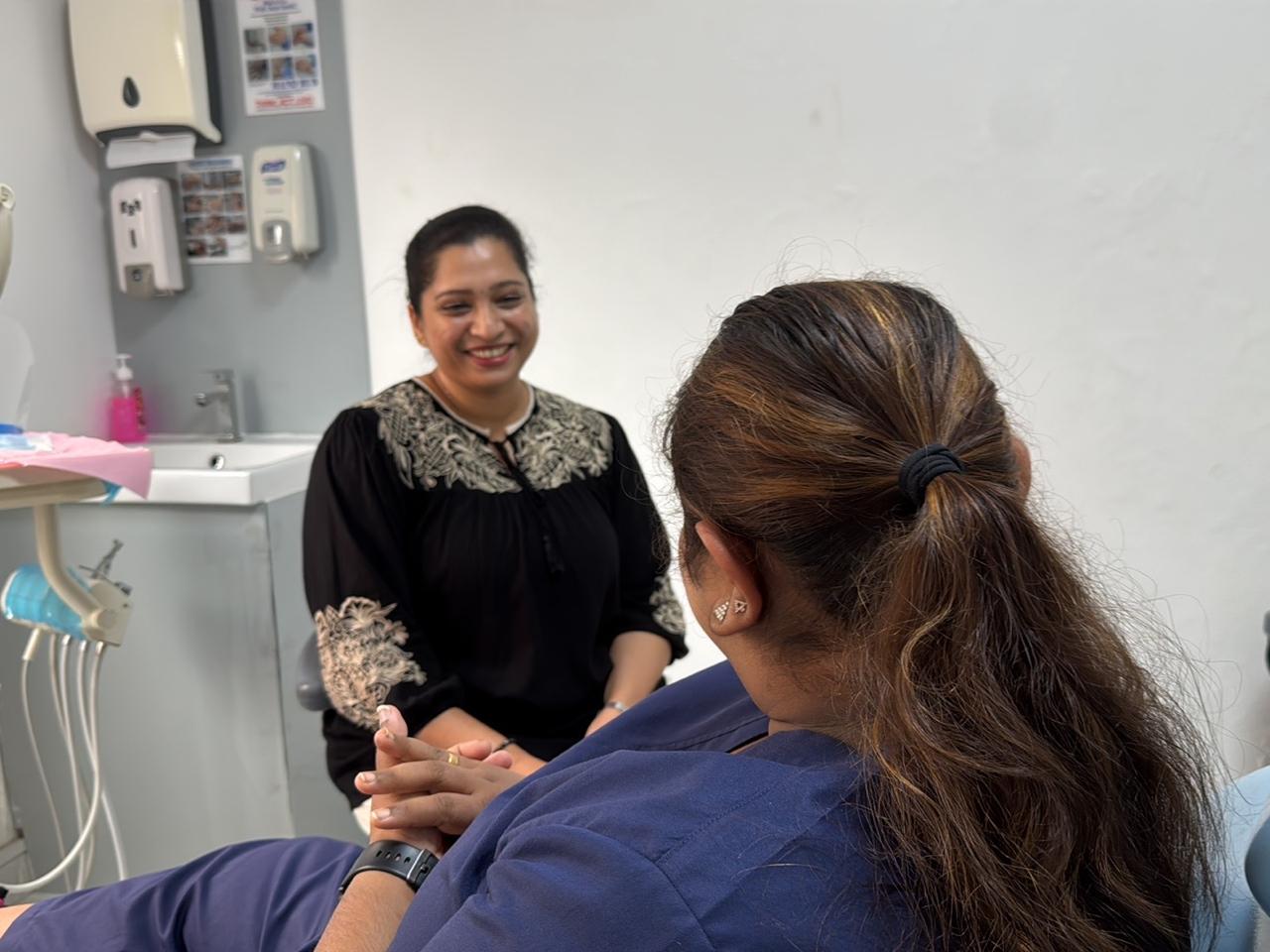 In a Best Dental Clinic treatment room, a woman in navy scrubs sits in a light-blue dental chair while another woman in a black embroidered blouse sits facing her and smiles; dental equipment, a sink and hand-hygiene posters are visible behind them